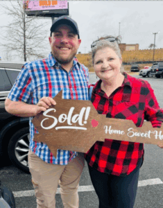 A smiling couple standing in a parking lot holding a wooden Sold sign Home Sweet Home, celebrating the successful sale of their home.